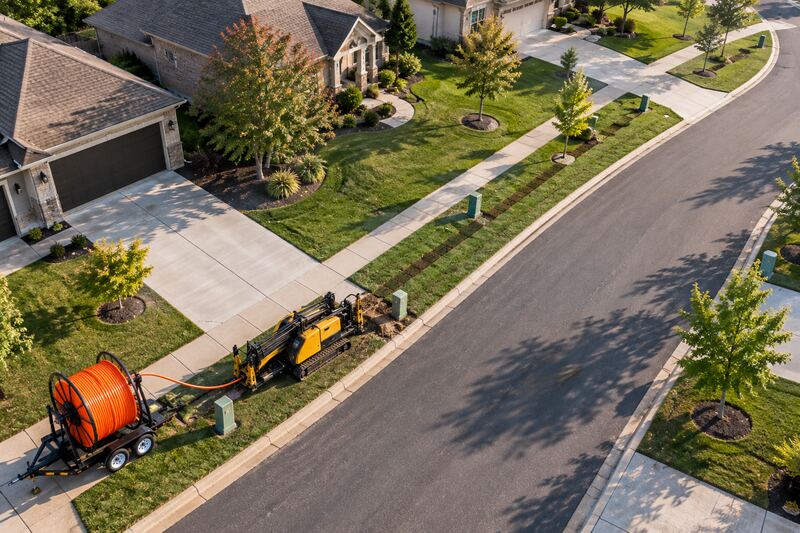 Drone view of a fiber-to-the-home build along a residential street: a restored strip of turf, green fiber pedestals, a reel of orange conduit and a compact drill.