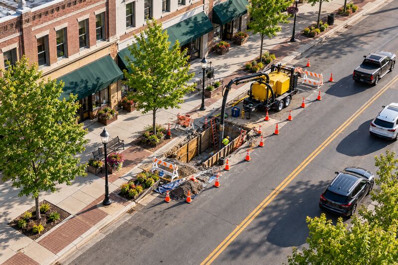 Drone view of a utility work zone on a main street, with an excavation, traffic cones, barricades and a trailer-mounted vacuum excavator beside storefronts.