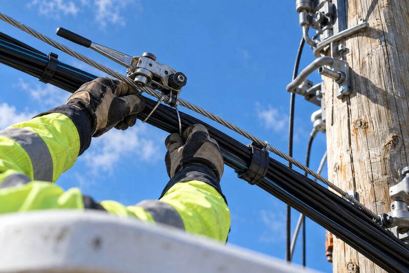 Gloved hands lashing black fiber optic cable to a steel support strand at the top of a wooden utility pole, under a blue sky.