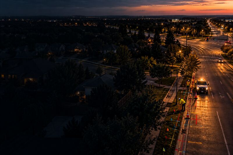 Aerial view of a suburban street at dusk, with a worker marking utility locate paint beside the curb next to a pickup and traffic cones.