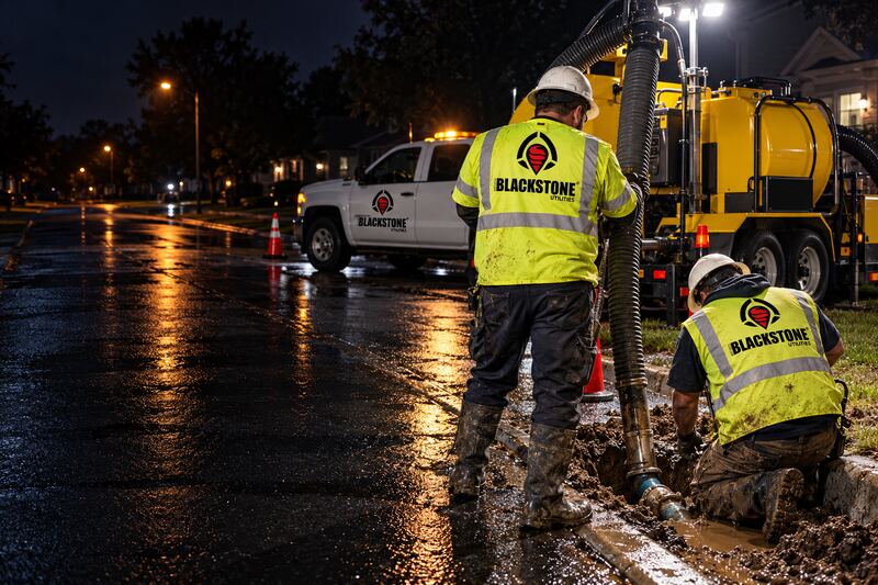 Two workers in Blackstone Utilities vests at a muddy excavation on a wet residential street at night, one holding a vacuum hose, with a yellow vacuum truck and a branded pickup with flashing lights.